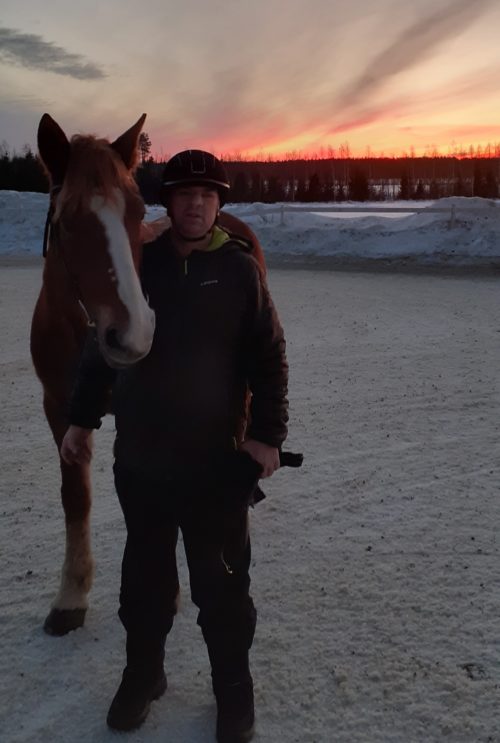Mies seisoo hevosen kanssa ilta-auringossa.