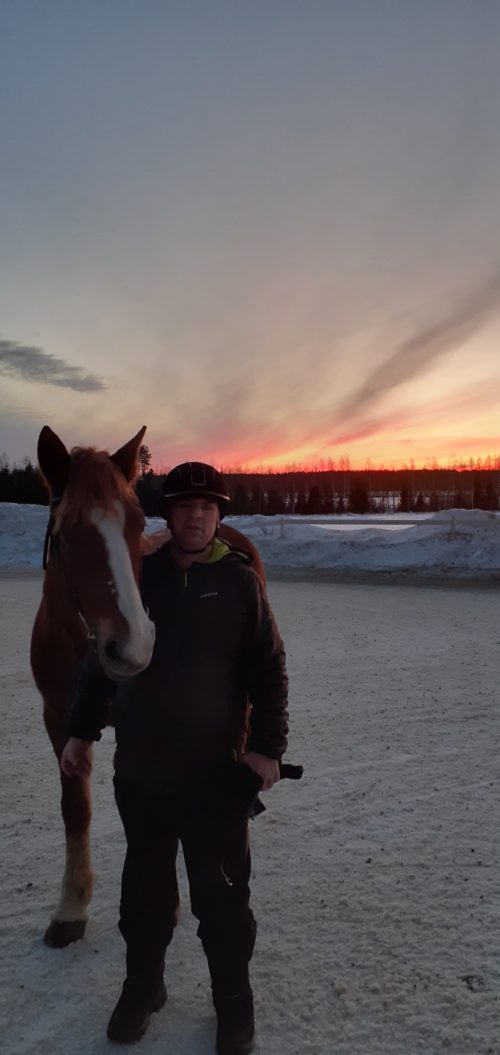 Mies seisoo hevosen vieressä ja taustalla auringonlasku.