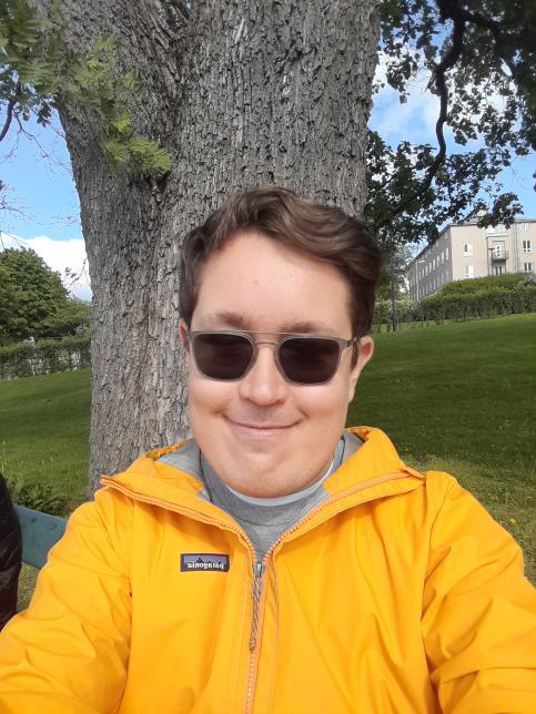 Mies hymyilee kameralle. Miehellä on aurinkolasit ja keltainen takki.
