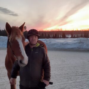 Mies ja suomenhevonen lumisella kentällä ja auringon laskussa.