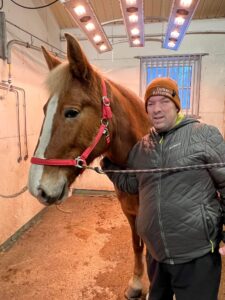Mies seisoo tallissa hevosen kanssa, katsoo kameraan ja hymyilee.