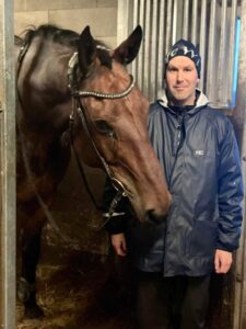 Mies seisoo ja hymyilee hevosen vieressä sadetakki päällä.