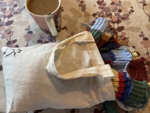 Kuppi kahvia ja kangaskassi, jossa on neuloksia. Kassiin on maalattu pieni, musta kissa.