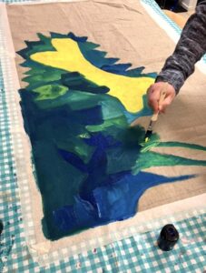 Nainen maalaa vihreitä, sinisiä ja keltaisia hahmoja kankaalle.