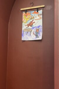 Vesivärityö roikkuu henkarissa ruskealla seinällä. Työssä on trampoliini, lintu, jänis ja kettu.