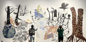 Seinällä iso maalaus, jossa puita juurinen ja monenlaisia hahmoja.
